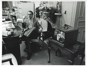 Mike Reed seated at a desk. There is a television screen with Carl Albert on it beside him., 1950-01-01 - 1976-12-31