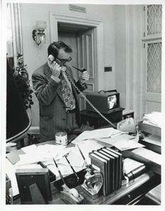 Mike Reed seated at a desk. There is a television screen with Carl Albert on it beside him., 1950-01-01 - 1976-12-31