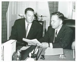 Carl Albert seated at a desk. He is looking at a document with another man., 1950-01-01 - 1976-12-31