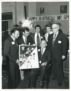Carl Albert seated in his new chair holding up his portrait of Oklahoma football. Several men are standing around him, including Tom Steed, 1976, 1976-05-10