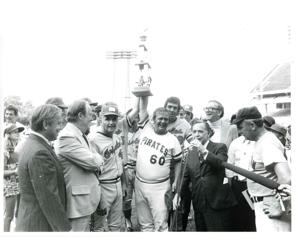 Carl Albert standing with several baseball players. Albert is speaking into a microphone and two baseball players are holding up a trophy. 1976, 1976-01-01 - 1976-12-31