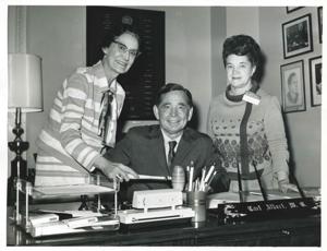 Carl Albert seated at his desk. Two unidentified women are standing behind him., 1950-01-01 - 1976-12-31