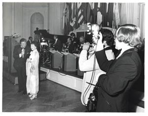 Mary Albert apparently being introduced at the American Revolution Bicentennial Ball in Oklahoma. She is standing beside a man with a microphone. A display of flags and an orchestra is shown behind them. October 02, 1976., 1976-10-02
