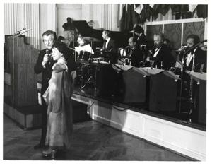 Mary Albert apparently being introduced at the American Revolution Bicentennial Ball in Oklahoma. She is standing beside a man with a microphone. A display of flags and an orchestra is shown behind them. October 02, 1976., 1976-10-02
