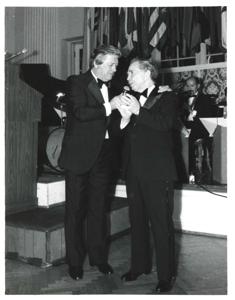 Mary Albert apparently being introduced at the American Revolution Bicentennial Ball in Oklahoma. She is standing beside a man with a microphone. A display of flags and an orchestra is shown behind them. October 02, 1976., 1976-10-02